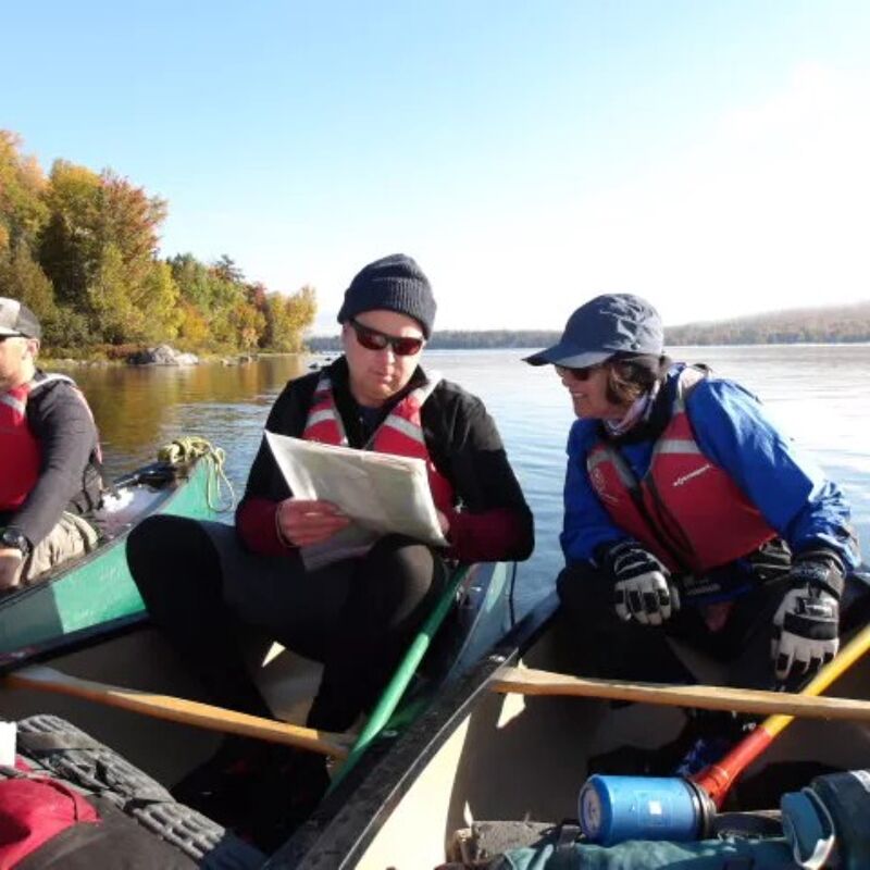 Two people in canoes are on a lake, possibly navigating. The person on the left is holding a map and wearing a black beanie, sunglasses, and a red life vest. The person on the right is wearing a baseball cap, sunglasses, and a blue jacket with a red life vest. Both are equipped with paddles, and the scenery includes trees and a clear sky.